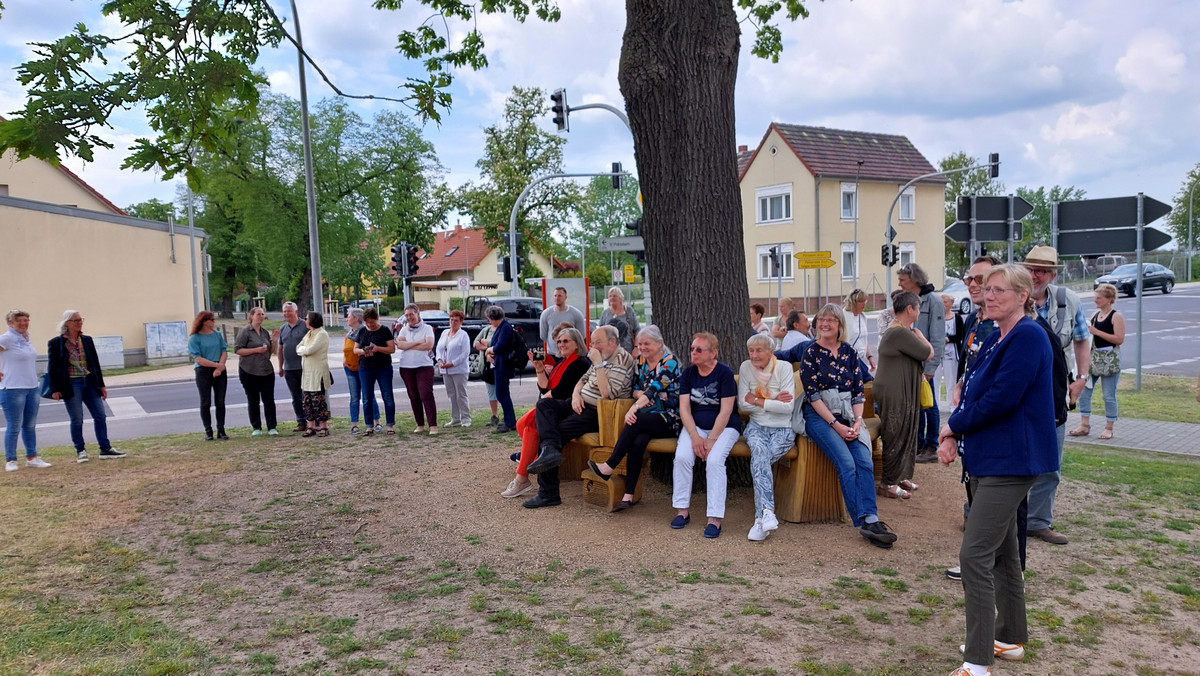 Eine Gruppe von Menschen sitzt auf einer Bank, die rund um einen Baum verläuft. Weitere Menschen stehen darum. - Klick öffnet Bildbetrachter