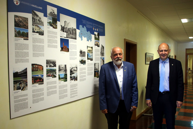 Zwei Männer stehen in einem Flur, an der Wand hängt eine Schautafel.