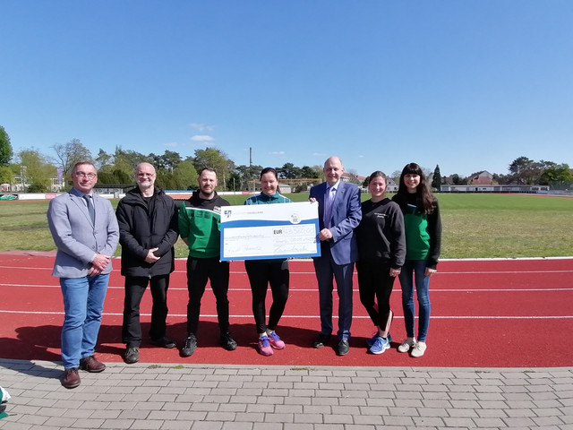 7 Personen stehen auf der Laufbahn im Premnitzer Stadion und die 3 in der Mitte halten den symbolischen Spendenscheck. - Klick öffnet Bildbetrachter