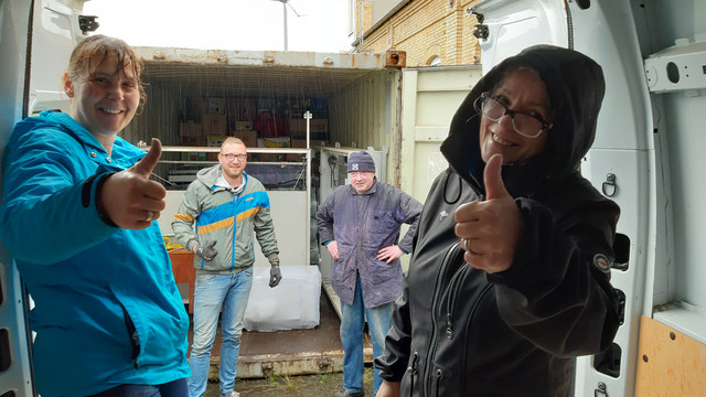 Mitglieder des Vereins Hand in Hand stehen am Container mit den eingeladenen Tafeln und schauen in die Kamera.