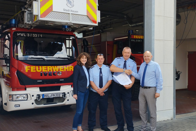Vier Personen stehen vor einer Feuerwehr-Halle, in der zwei Feuerwehren stehen - Klick öffnet Bildbetrachter