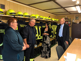 Vier Personen stehen in einem Raum und unterhalten sich. Im Hintergrund hängen Feuerwehruniformen.