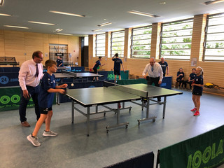 Vier Personen, zwei Kinder und zwei Erwachsene, stehen in einer Doppelformation an einer Tischtennisplatte.