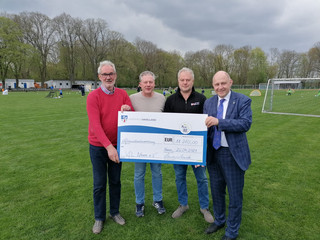 Vier Personen stehen auf dem Fußballplatz und halten den symbolischen Spendenscheck. - Klick öffnet Bildbetrachter