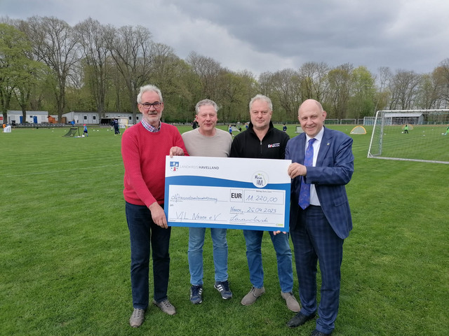 Vier Personen stehen auf dem Fußballplatz und halten den symbolischen Spendenscheck. - Klick öffnet Bildbetrachter