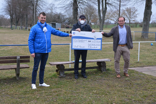 Drei Männer stehen an einem Sportplatz und halten einen symbolischen Scheck in den Händen.