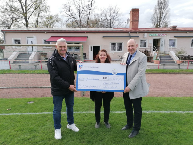 Drei Personen stehen auf dem Fußballplatz und halten den symbolischen Spendenscheck. - Klick öffnet Bildbetrachter