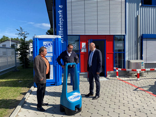 Drei Männer stehen vor einer Dixi-Toilette und unterhalten sich.