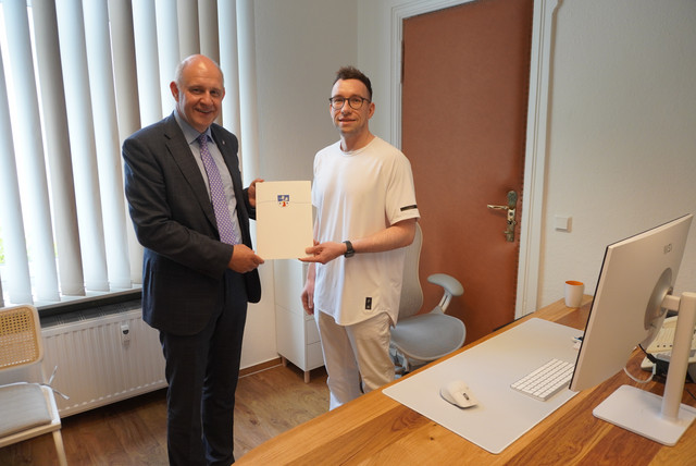 Der Landrat und der Arzt stehen in einem Behandlungszimmer und halten zwischen sich eine Mappe mit Landkreiswappen - Klick öffnet Bildbetrachter