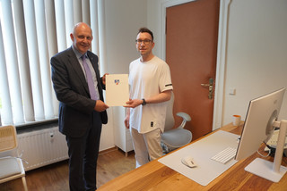 Der Landrat und der Arzt stehen in einem Behandlungszimmer und halten zwischen sich eine Mappe mit Landkreiswappen - Klick öffnet Bildbetrachter