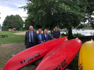 Vier Personen stehen hinter drei auf dem Kopf liegenden Kanu-Booten. Im Hintergund sieht man Wasser.
