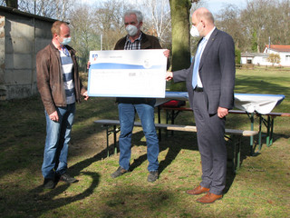 Drei Männer stehen an einem Sportplatz und halten einen symbolischen Scheck in den Händen.