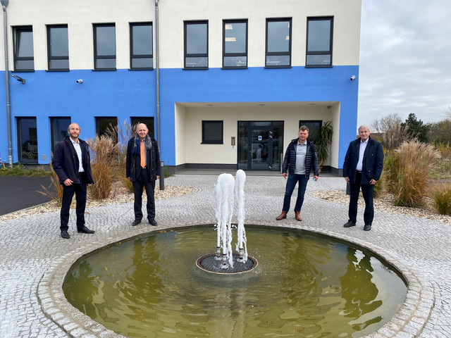 Vier Männer stehen vor einem Gebäude, verteilt um ein Wasserspiel.