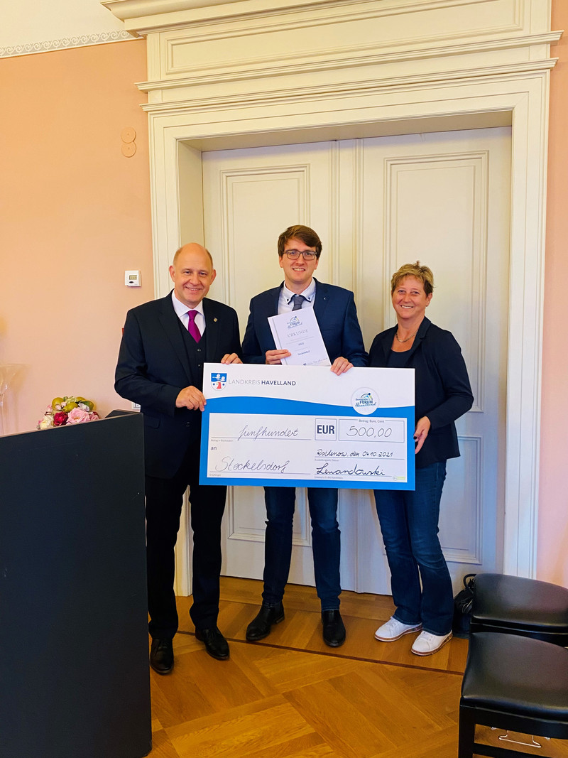 Drei Personen stehen neben einander, die Person in der Mitte hält einen symbolischen Siegerscheck.