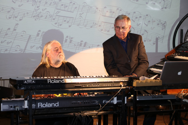 Wolfgang Fuchs sitzt links und spielt auf dem Keyboard. Rechts daneben sitzt Dietrich Joecks und beobachtet ihn.