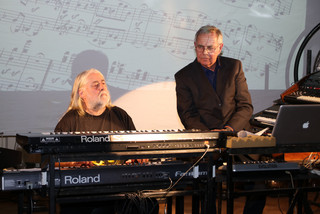 Wolfgang Fuchs sitzt links und spielt auf dem Keyboard. Rechts daneben sitzt Dietrich Joecks und beobachtet ihn.