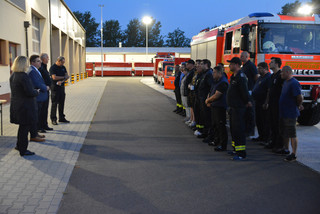 Vier Personen stehen vor einer Gruppe von Feuerwehrleuten, im Hintergrund stehen verschiedene Feuerwehrfahrzeuge.