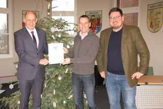 Drei Männer stehen an einem Weihnachtsbaum, zwei halten eine Dokumentenmappe zwischen sich.