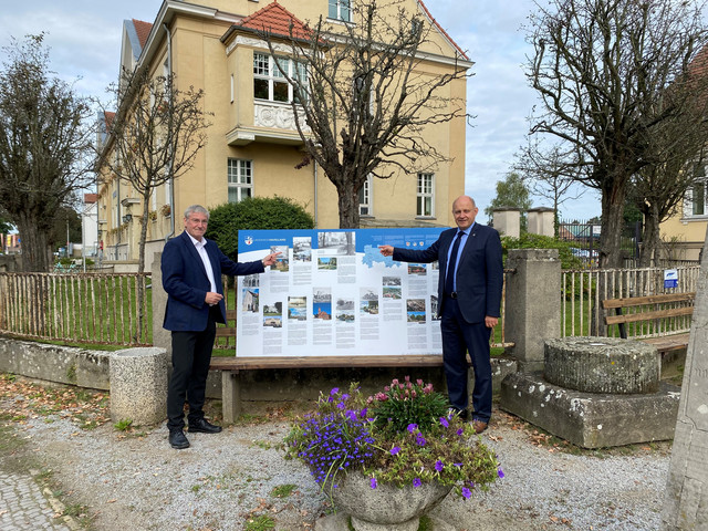 Zwei Männer stehen vor einem Gebäude, zwischen ihnen steht auf einer Bank die Schautafel.