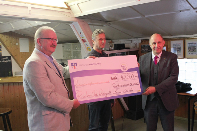 Drei Männer stehen in einem Raum und halten gemeinsam einen großen Scheck in den Händen.