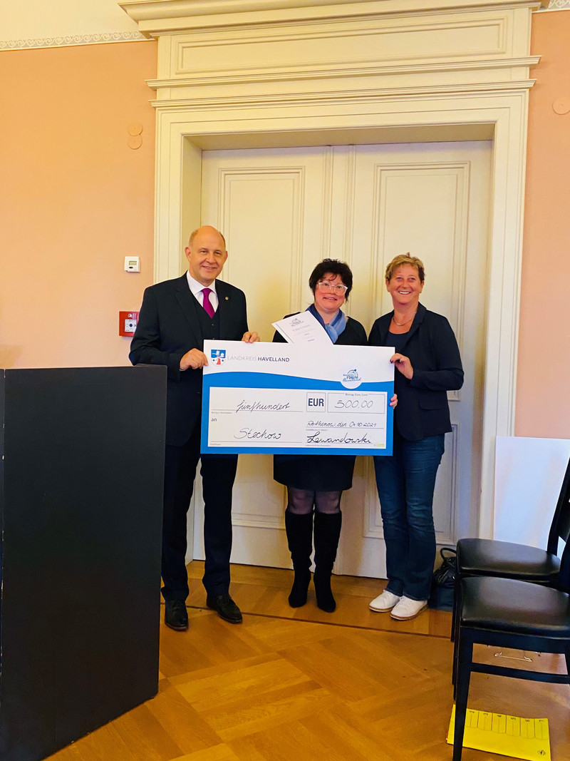 Drei Personen stehen neben einander, die Person in der Mitte hält einen symbolischen Siegerscheck.