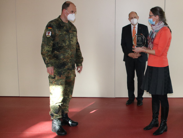 Karen Heinrich steht rechts und überreicht Oberstleutnant Klitscher, der links steht, einen Glaspokal. Im Hintergrund steht Landrat Roger Lewandowski.
