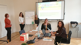 Fünf Frauen sitzen und stehen verteilt in einem Raum.