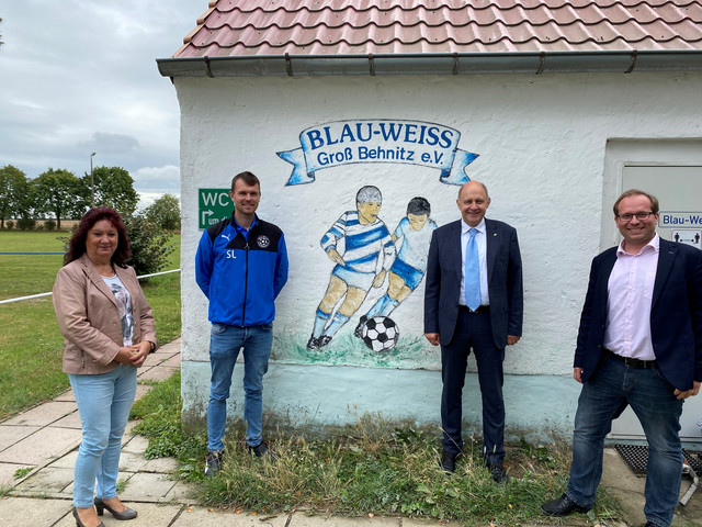 Vier Personen stehen vor einem Vereinsgebäude und schauen in die Kamera.