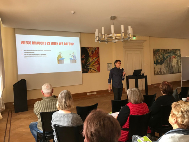 Klick öffnet Bildbetrachter; ein Mann steht vor einer Gruppe Menschen