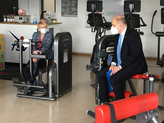 Landrat Lewandowski und Birgit Faber sitzen auf Sportgeräten in einem Fitness-Raum und unterhalten sich..