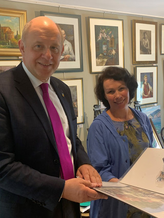 Zwei Personen schauen in die Kamera, im Hintergrund hängen Bilder an der Wand - Klick öffnet Bildbetrachter