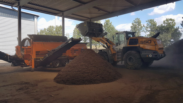 Ein Bagger schüttet Abfall in die Kompostieranlage, davor liegt ein großer Haufen Kompost - Klick öffnet Bildbetrachter