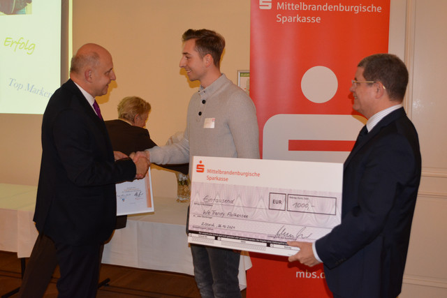 Ein Mann schüttelt die Hand eines anderen Mannes. Daneben steht ein weiterer Mann mit einem großen Spendenscheck.