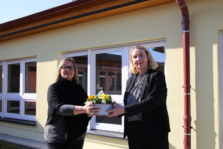 Zwei Frauen stehen vor einem Gebäude und halten zwischen sich eine Schale mit Blumen.