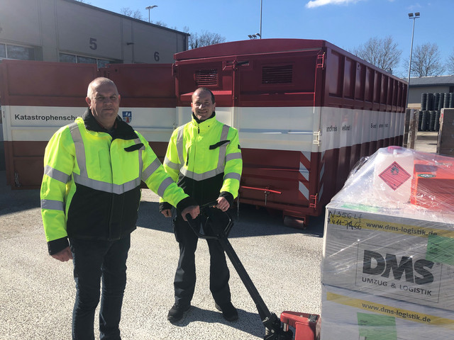 Zwei Männer stehen vor einem Container. Sie bedienen einen Hubwagen mit Materialien.