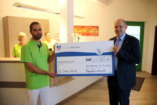 Dr. Wilke und Landrat Lewandowski stehen in der Arztpraxis und halten einen symbolischen Scheck zwischen sich.