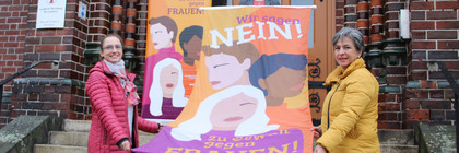 Drei Frauen stehen auf einer Treppe und halten eine orange Fahne mit der Aufschrift "Wir sagen Nein! zu Gewalt gegen Frauen".