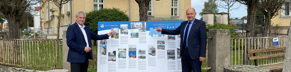 Zwei Männer stehen vor einem Gebäude, zwischen ihnen steht auf einer Bank die Schautafel.