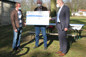 Drei Männer stehen an einem Sportplatz und halten einen symbolischen Scheck in den Händen.