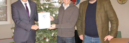 Drei Männer stehen an einem Weihnachtsbaum, zwei halten eine Dokumentenmappe zwischen sich.