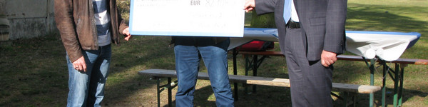 Drei Männer stehen an einem Sportplatz und halten einen symbolischen Scheck in den Händen.