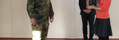 Karen Heinrich steht rechts und überreicht Oberstleutnant Klitscher, der links steht, einen Glaspokal. Im Hintergrund steht Landrat Roger Lewandowski.