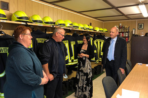 Vier Personen stehen in einem Raum und unterhalten sich. Im Hintergrund hängen Feuerwehruniformen.