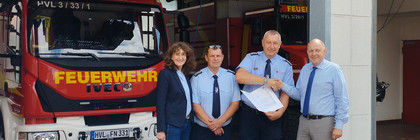 Vier Personen stehen vor einer Feuerwehr-Halle, in der zwei Feuerwehren stehen - Klick öffnet Bildbetrachter