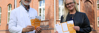 Zwei Personen stehen vor einem Backsteingebäude und halten Fyler in den Händen.