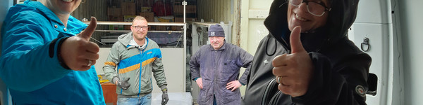 Mitglieder des Vereins Hand in Hand stehen am Container mit den eingeladenen Tafeln und schauen in die Kamera.