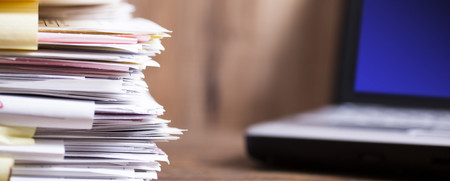 Large stack of files, paperwork.  Desk, office, computer.