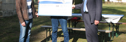 Drei Männer stehen an einem Sportplatz und halten einen symbolischen Scheck in den Händen.