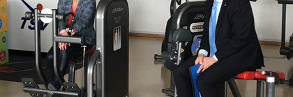 Landrat Lewandowski und Birgit Faber sitzen auf Sportgeräten in einem Fitness-Raum und unterhalten sich..