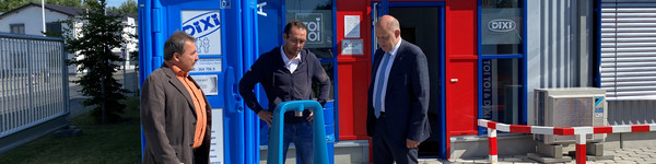 Drei Männer stehen vor einer Dixi-Toilette und unterhalten sich.